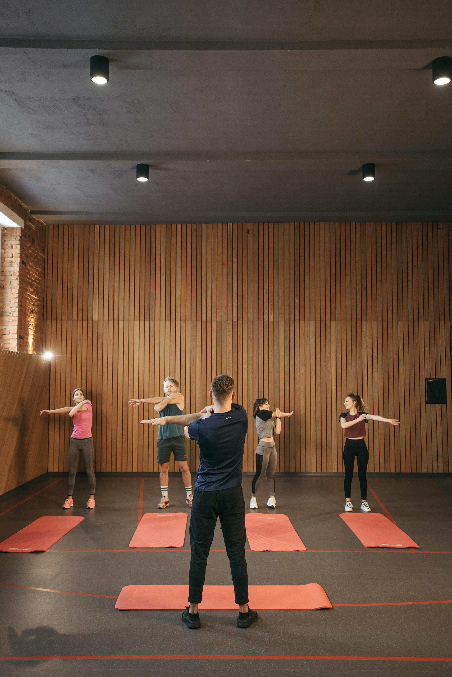 A diverse group of adults stretching together in an indoor fitness class, led by an instructor.